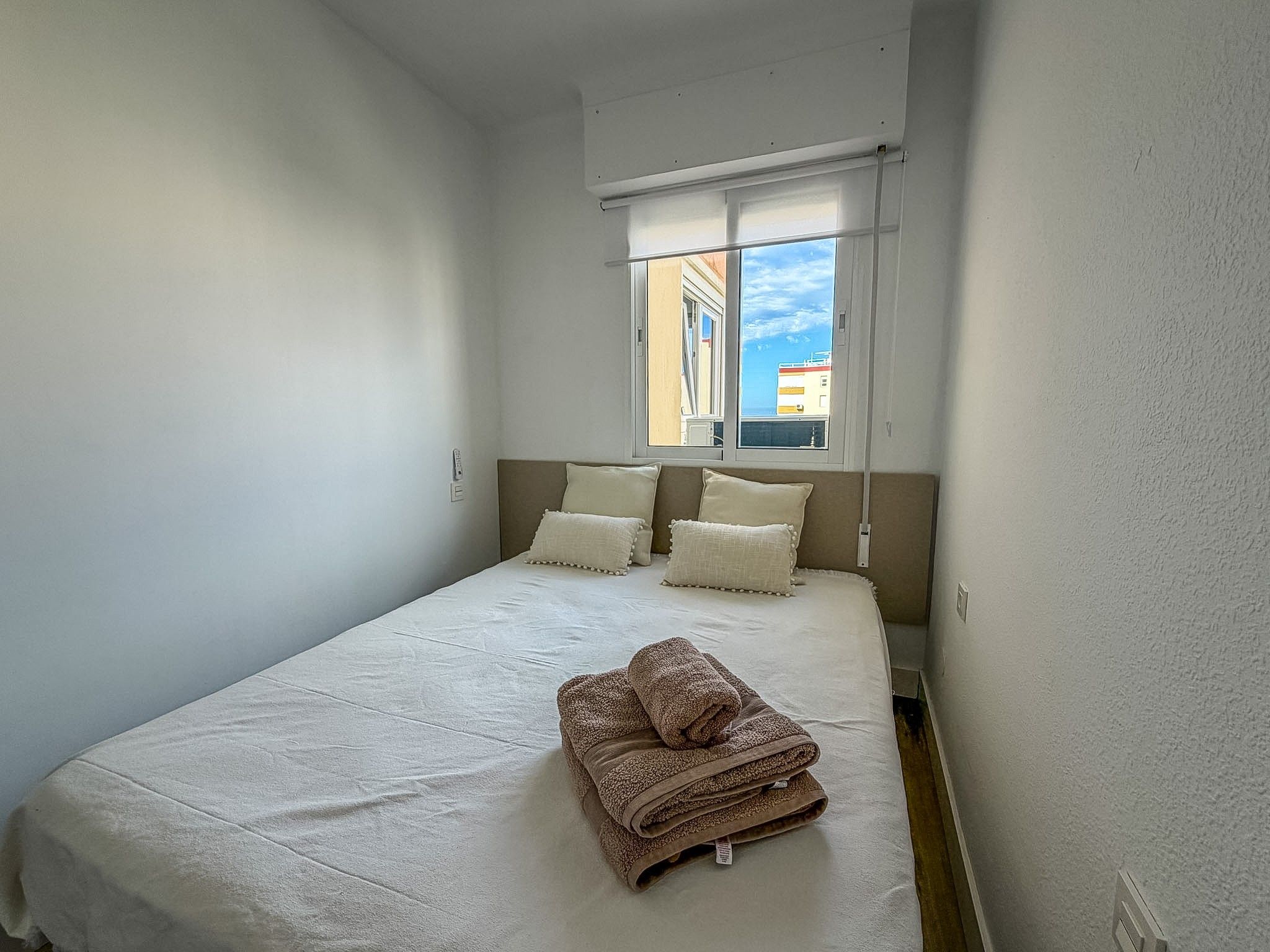 A single-bed room with a window, white walls, and a neatly made bed with pillows and a folded blanket.