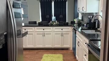 A kitchen with white cabinets, a stainless steel refrigerator, a green rug, and a window with dark curtains.