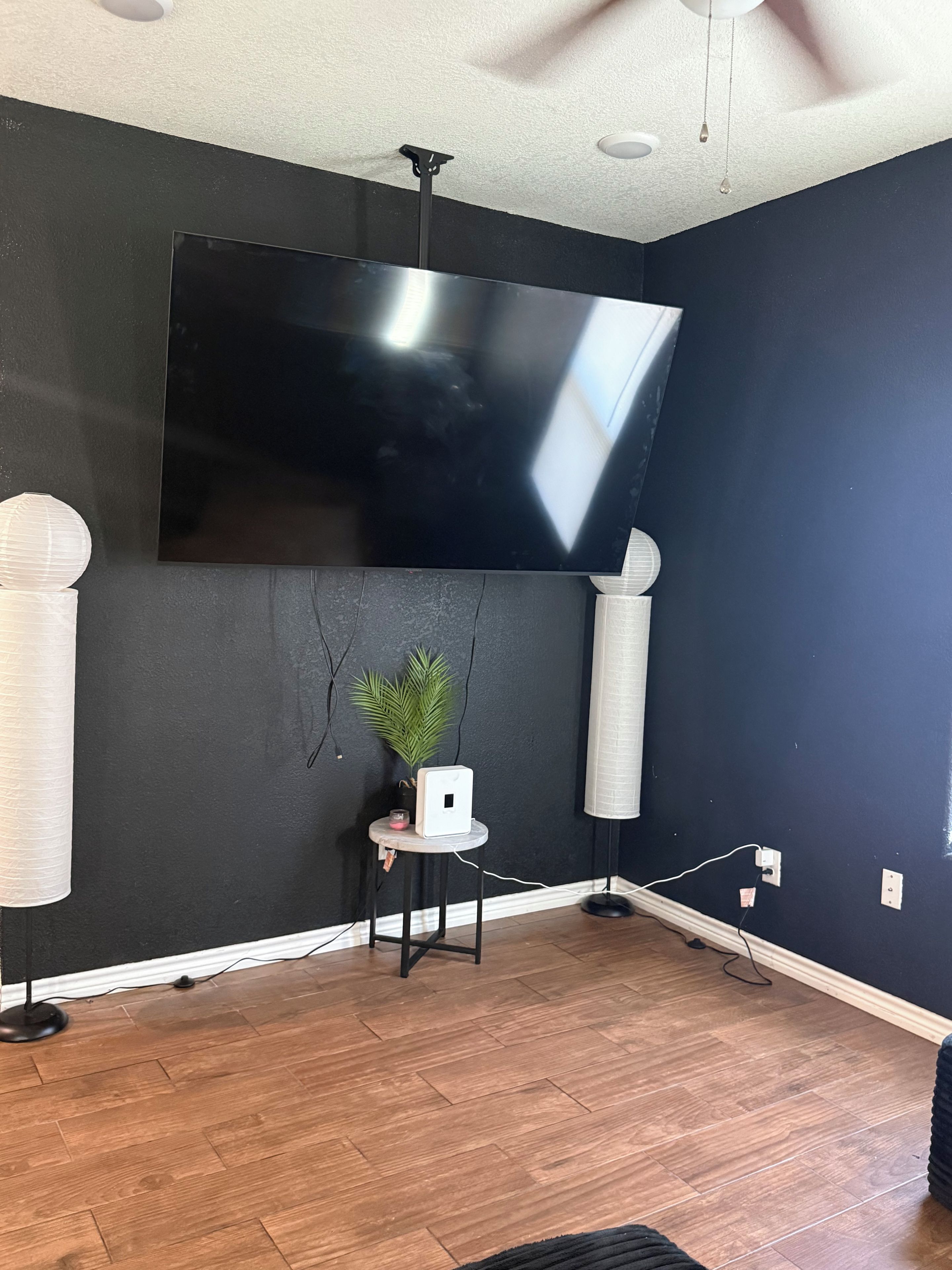 A room with a large flat-screen TV mounted on the wall, a small table with a plant, and a ceiling fan.