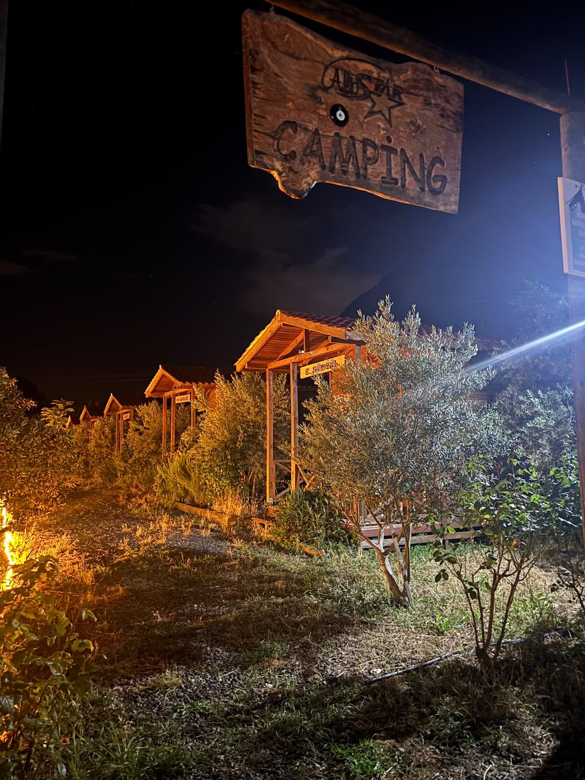 A wooden sign with 'CAMPING' written on it, surrounded by a rustic outdoor setting with wooden structures and vegetation.