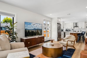 A modern living room with a large flat-screen TV, a wooden cabinet, a round coffee table, and a sofa. - South Campus Modern - Upscale & Walkable To Uo, Park & Shops 3 Bedroom Home (Eugene)