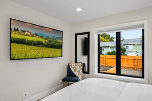 A bedroom with a large window, a bed, a chair, a painting, and a view of a house and fence. - South Campus Modern - Upscale & Walkable To Uo, Park & Shops 3 Bedroom Home (Eugene)