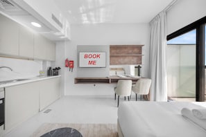 Una habitación de hotel moderna con área de cocina, cama con un edredón blanco impecable, un televisor montado mostrando 'BOOK ME' y un ventanal con cortinas.
