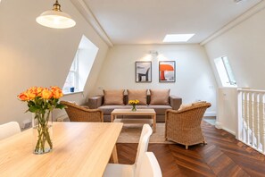 A modern living room with a sofa, two armchairs, a coffee table, and a dining table. There are two wall-mounted light fixtures and a window with a view. - Kensington 2-bed Mews House Next to Holland Park (London)