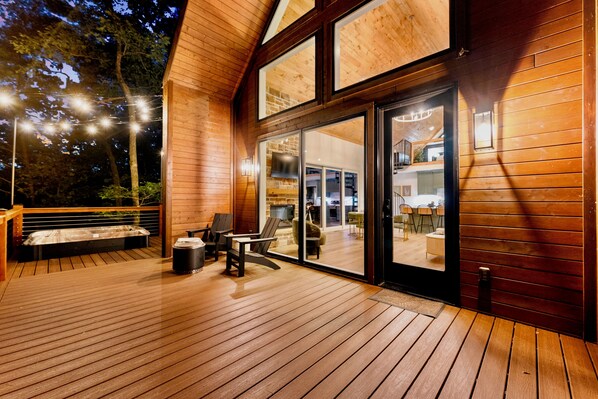 Una terraza de madera con mobiliario exterior y un amplio ventanal corredera que da a un espacio interior.