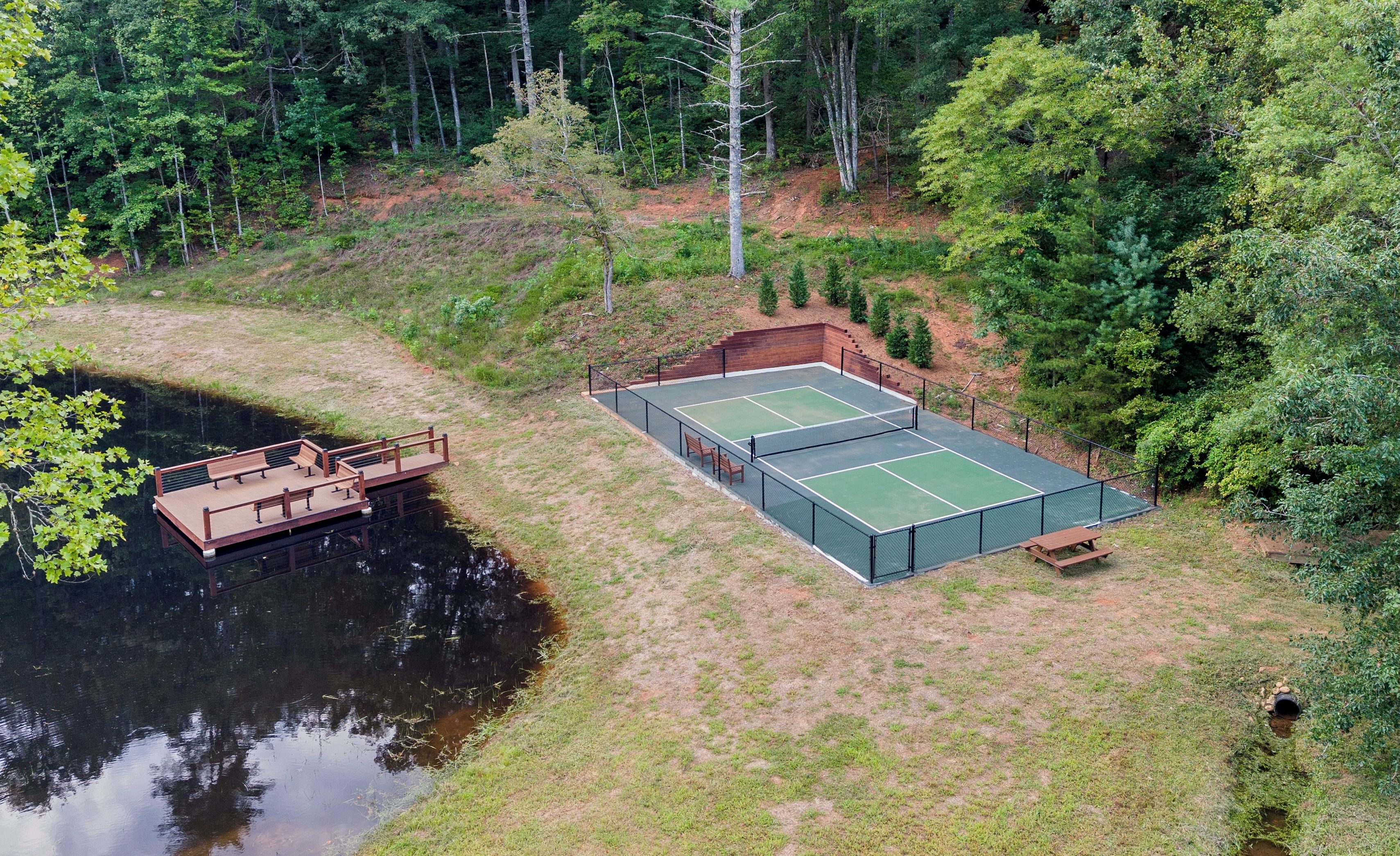 An outdoor tennis court with a wooden deck and benches, surrounded by trees and a body of water.