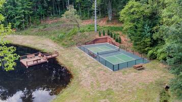 An outdoor tennis court with a wooden deck and benches, surrounded by trees and a body of water.