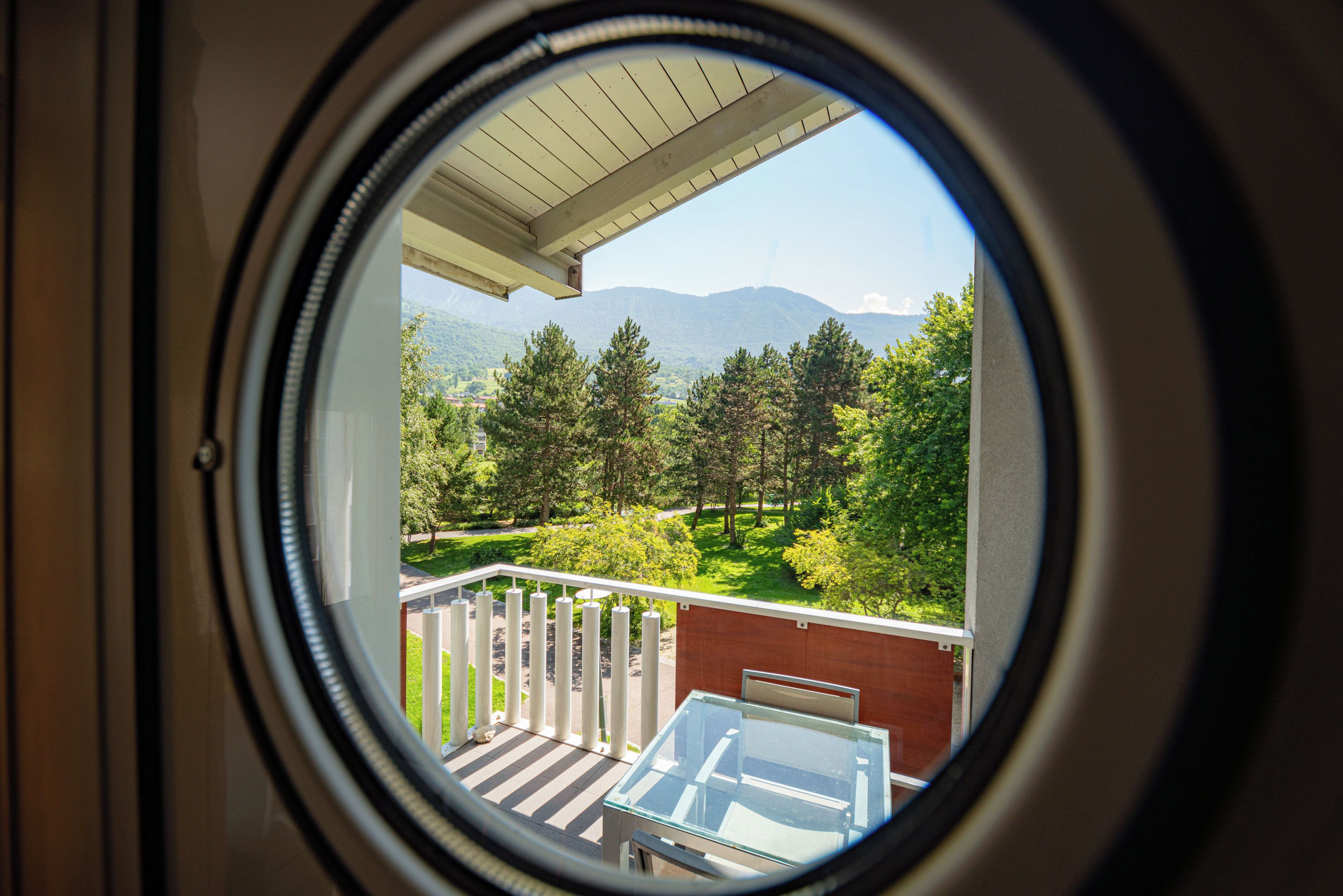 Une fenêtre circulaire donnant sur un balcon, une table en verre et un paysage montagneux.