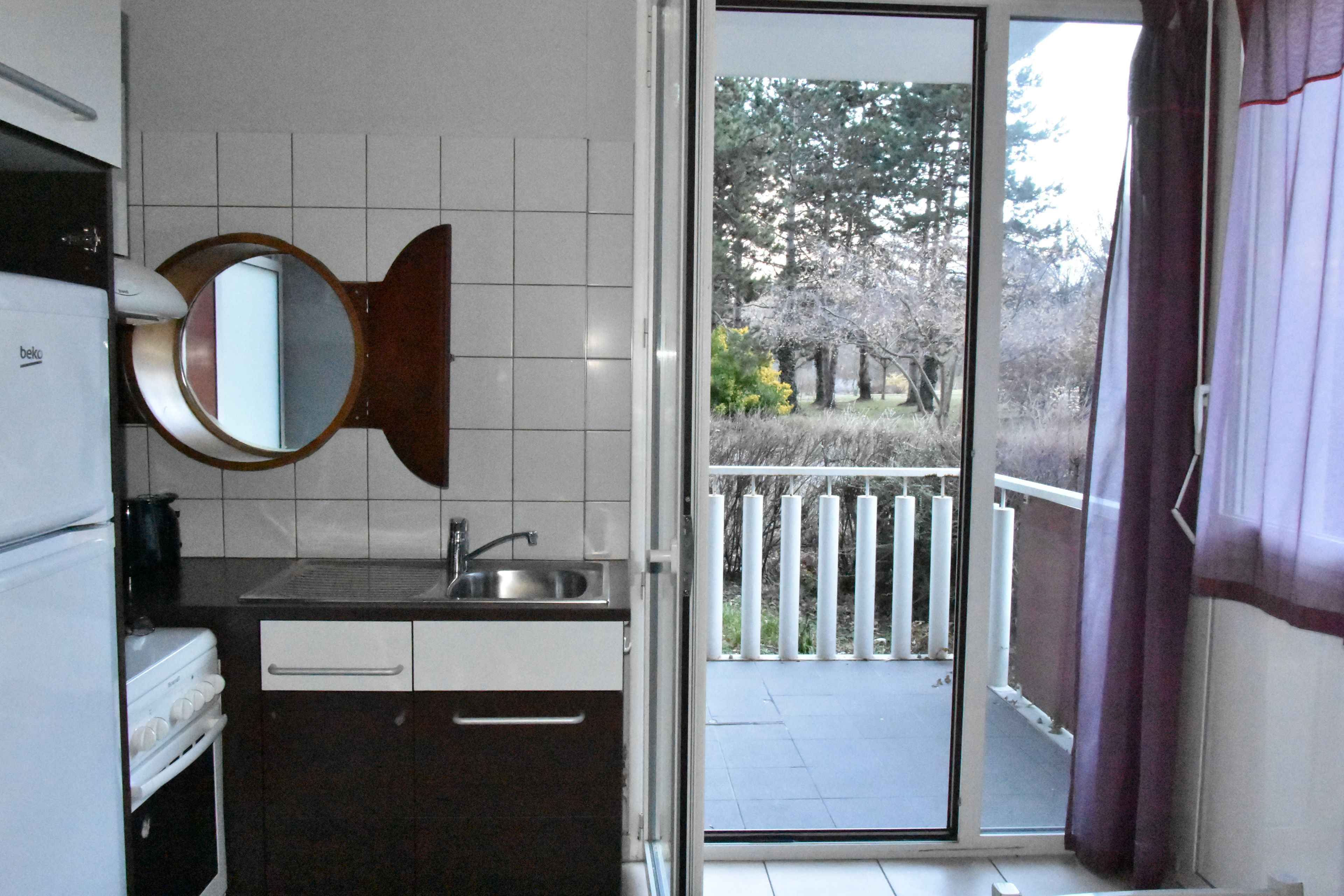 A compact kitchen with a sink, a mirror, and a window leading to a balcony with a view of trees.