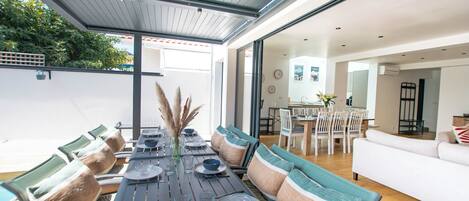 An outdoor dining area with a long table set for six, surrounded by chairs with patterned cushions.