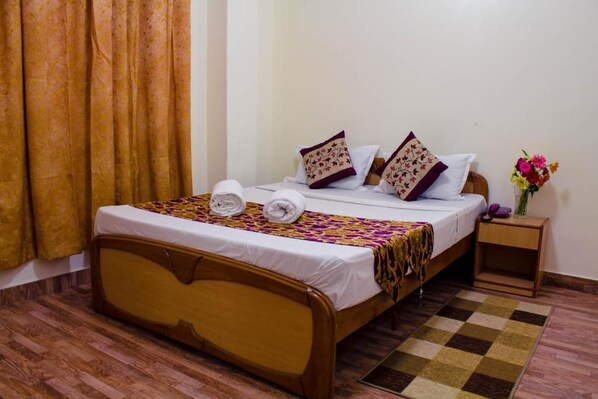 Cama bem arrumada com colcha estampada e almofadas, mesa de cabeceira de madeira com um vaso de flores, e tapete xadrez sobre piso de madeira.