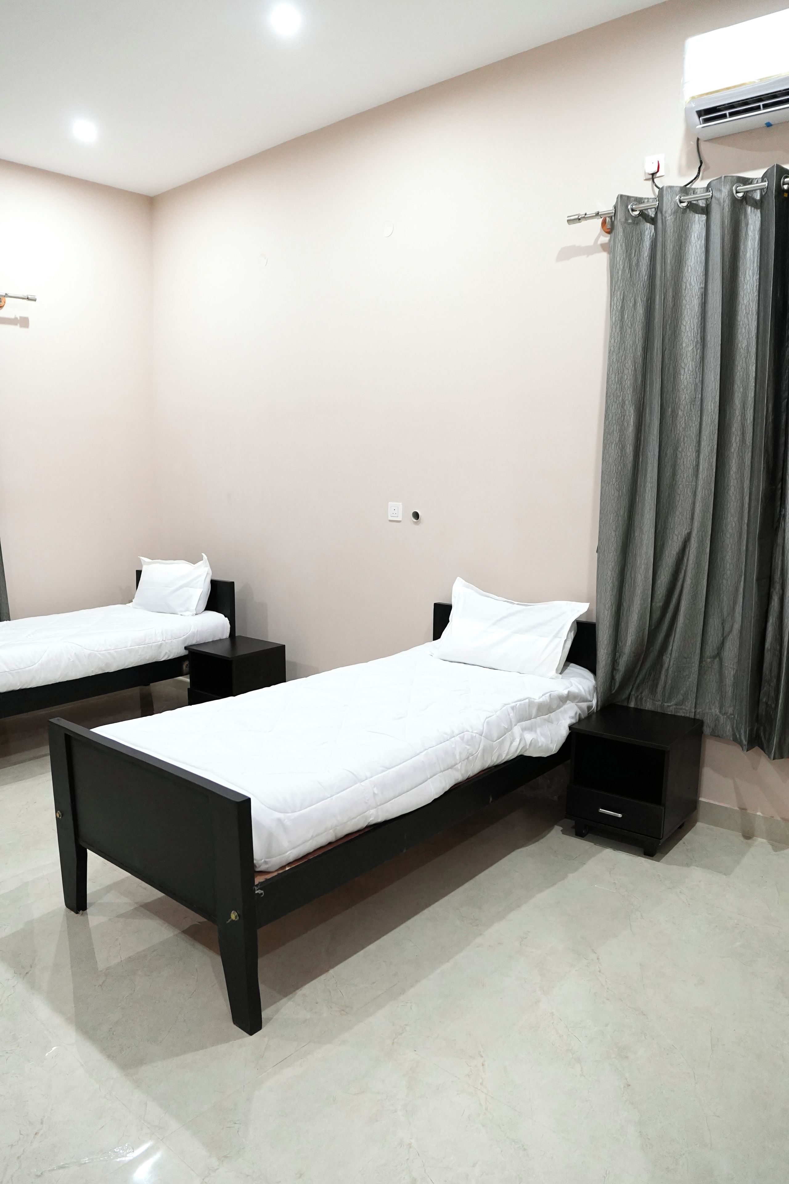 A room with two single beds, each with a nightstand, a wall socket, and a curtain.