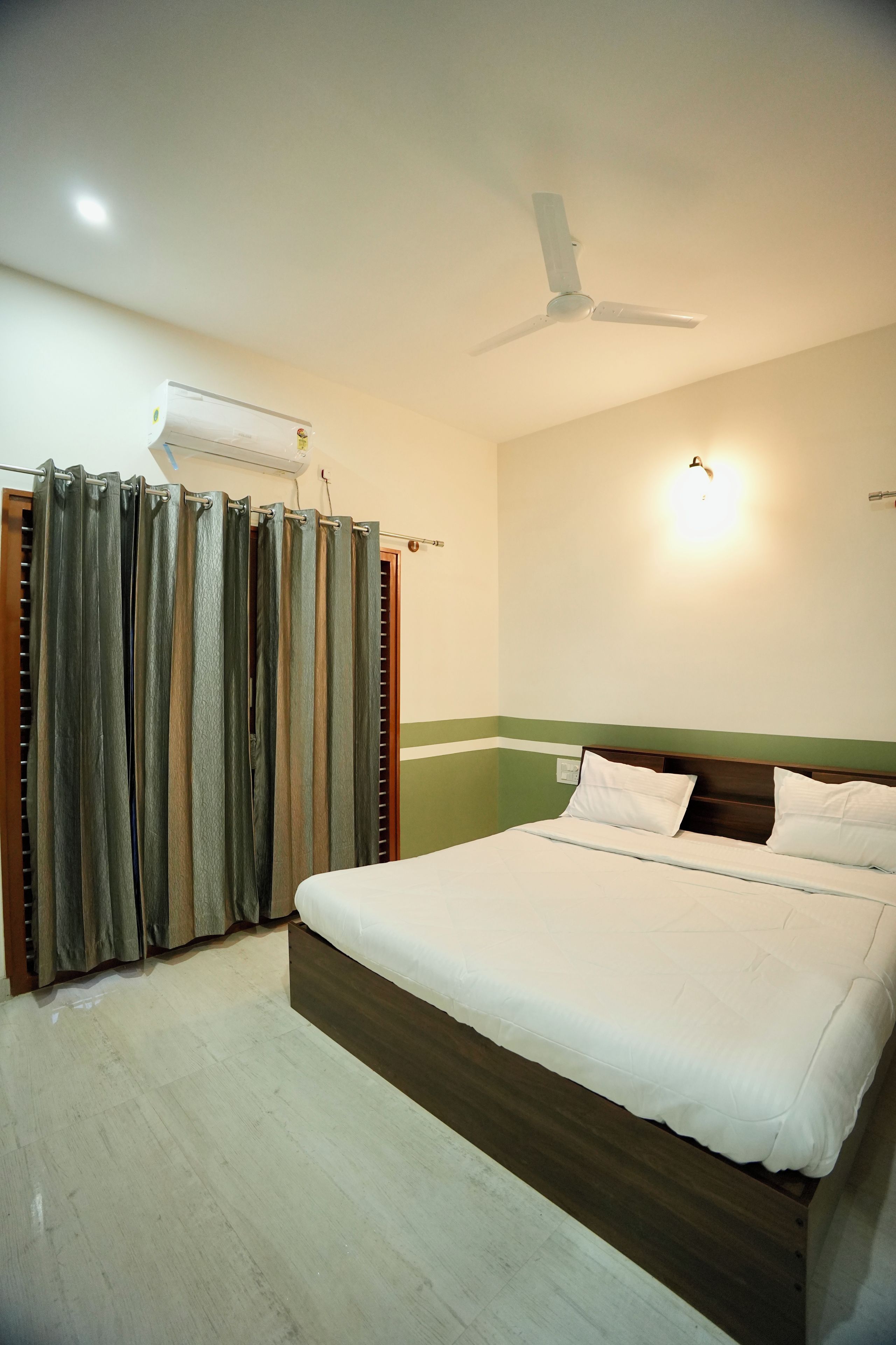 A hotel room with a bed, curtains, an air conditioning unit, a ceiling fan, and a wall-mounted light fixture.