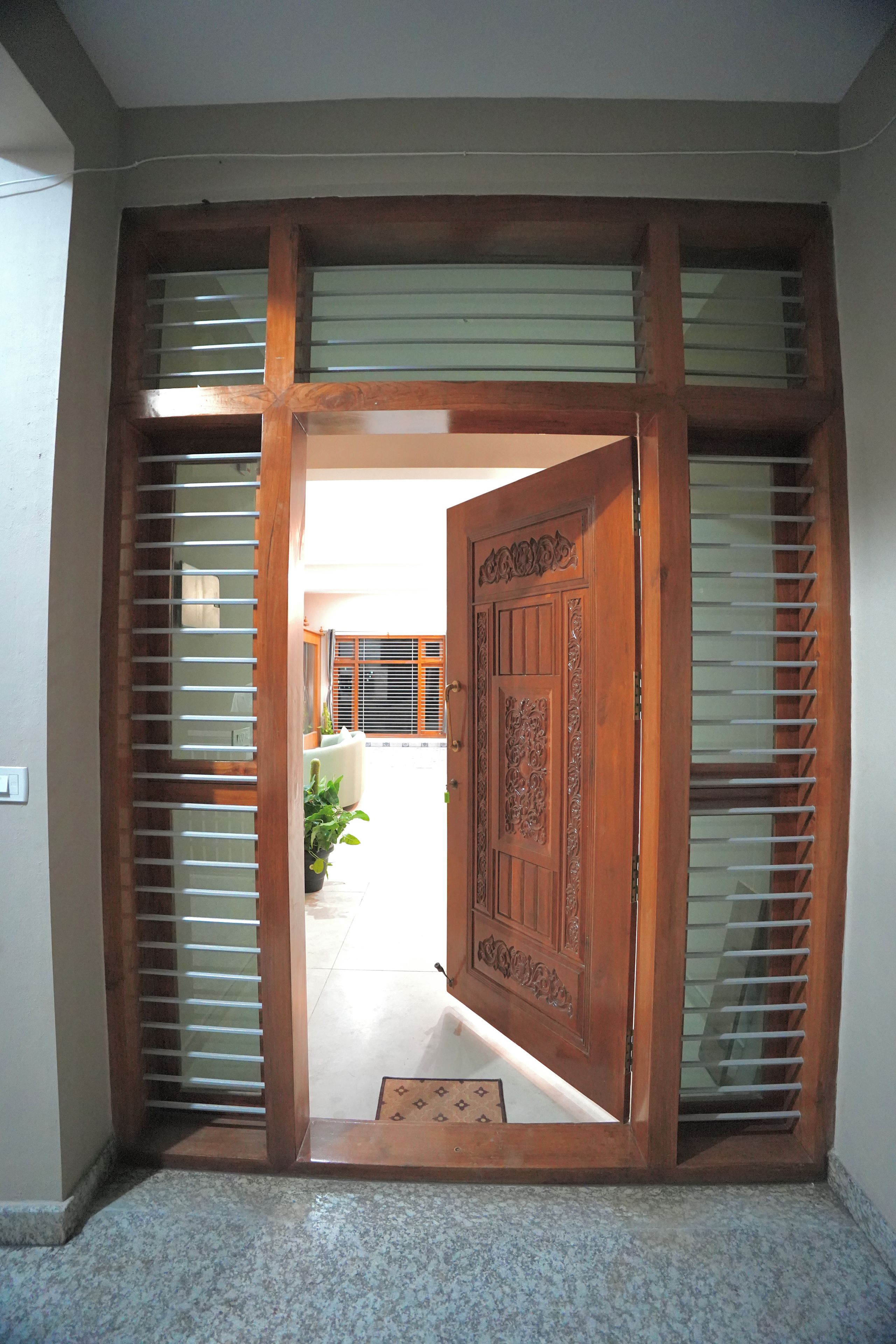 A wooden door with intricate carvings, flanked by vertical blinds, leading to a tiled corridor.