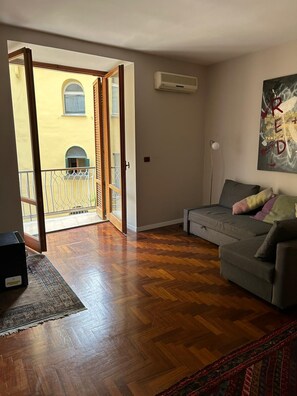 Ein offener Wohnbereich mit grauem Sofa, Holzboden, einer Schiebetür zum Außenbalkon und einer Wand-Klimaanlage.