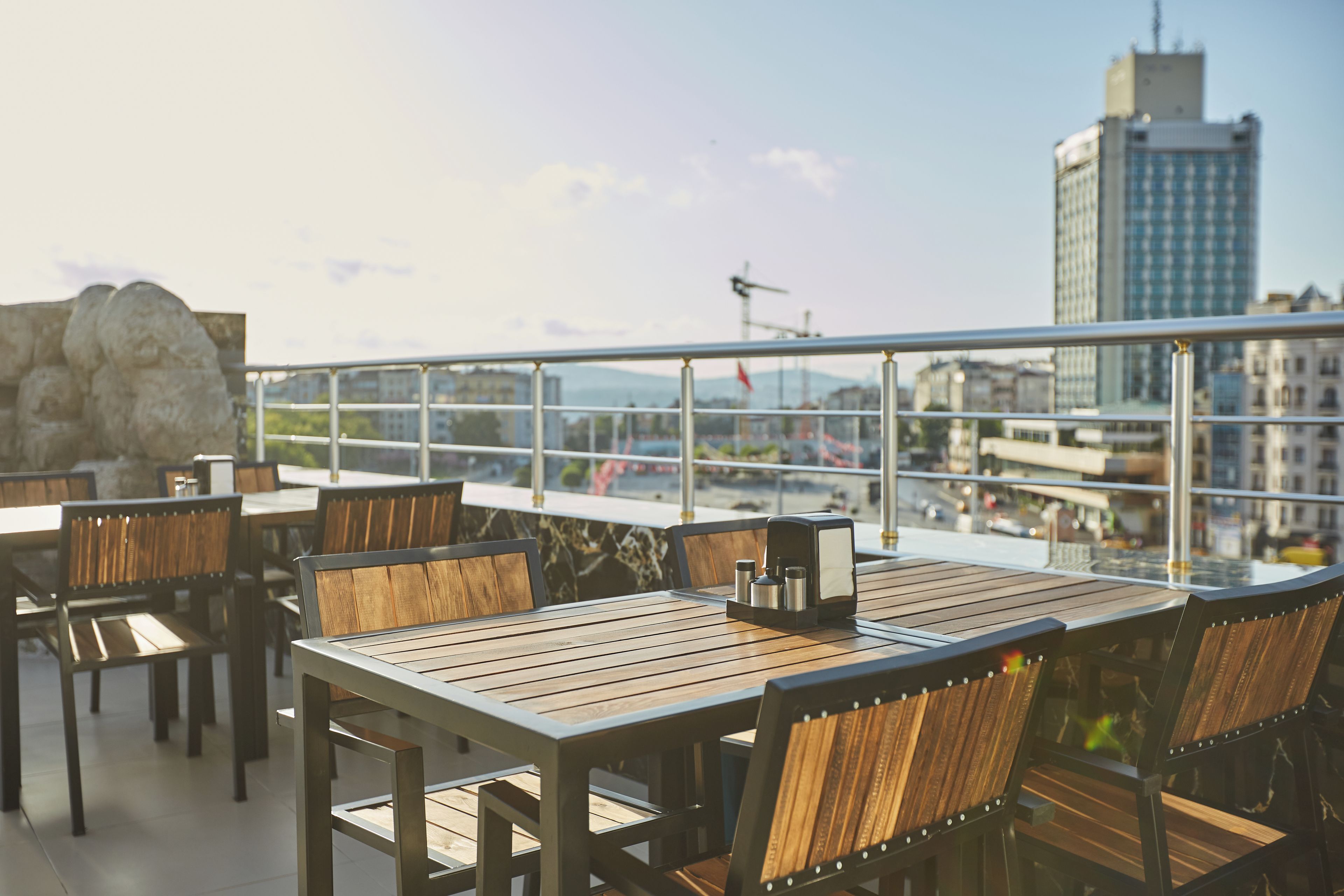An outdoor seating area with wooden tables and chairs, a stone wall, and a view of a cityscape with buildings and a crane.
