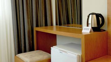 Camera d'albergo compatta con una scrivania in legno, un piccolo frigorifero, una panca imbottita e un bollitore sulla scrivania.