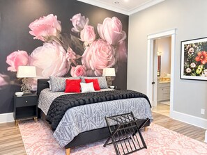 Ein Schlafzimmer mit einer großen Blumenmalerei an der Wand, einem Bett mit gemusterten Betttextilien, Nachttischen mit Lampen und Blick ins Badezimmer.