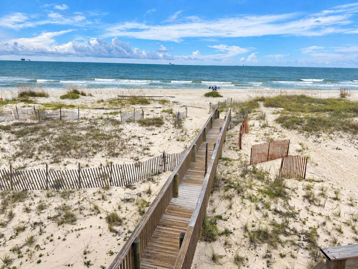 Une promenade en bois qui mène à une plage bordée de dunes de sable et d’une végétation clairsemée.