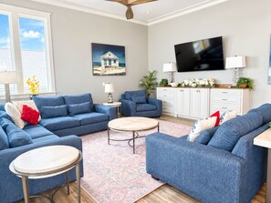A living room with blue sofas, a coffee table, a TV, and a painting on the wall. - Coco Bella – Gulf Front Luxury | Private Pool | Sleeps 8 (Fort Morgan)