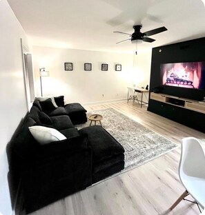 Ein modernes Wohnzimmer mit einer schwarzen Couch, einem weißen Sessel, einem Fernseher und einem Deckenventilator.