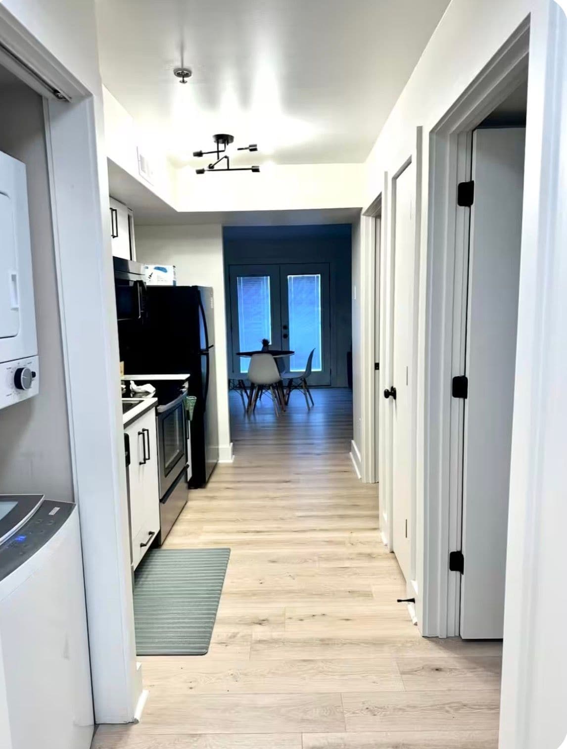 A narrow hallway with a kitchen on one side, a dining area with a table and chairs, and a door leading to another room.