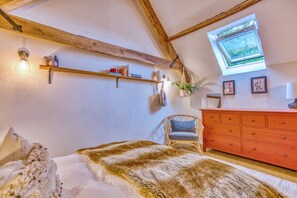 Ein Schlafzimmer mit schrägem Dach, Holzbalken, einem Bett mit Felldecke, einem Korbstuhl, einer Kommode und einem Fenster mit Blick.