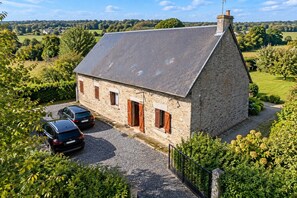 Une maison en pierre avec un toit en pente, une allée gravillonnée et deux voitures garées.