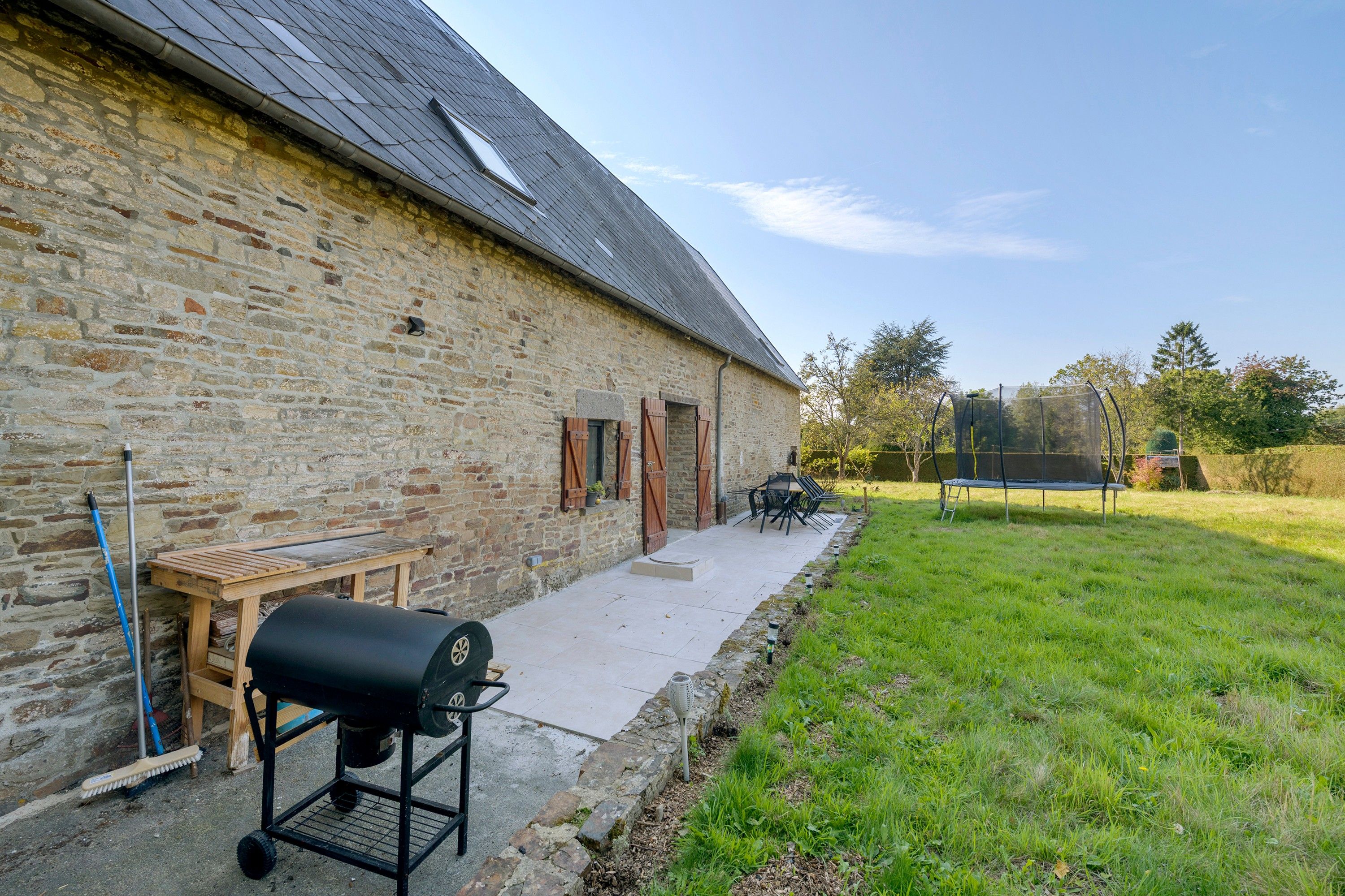 Een gebouw van steen met een schuin dak, een barbecue en een buitentafel.