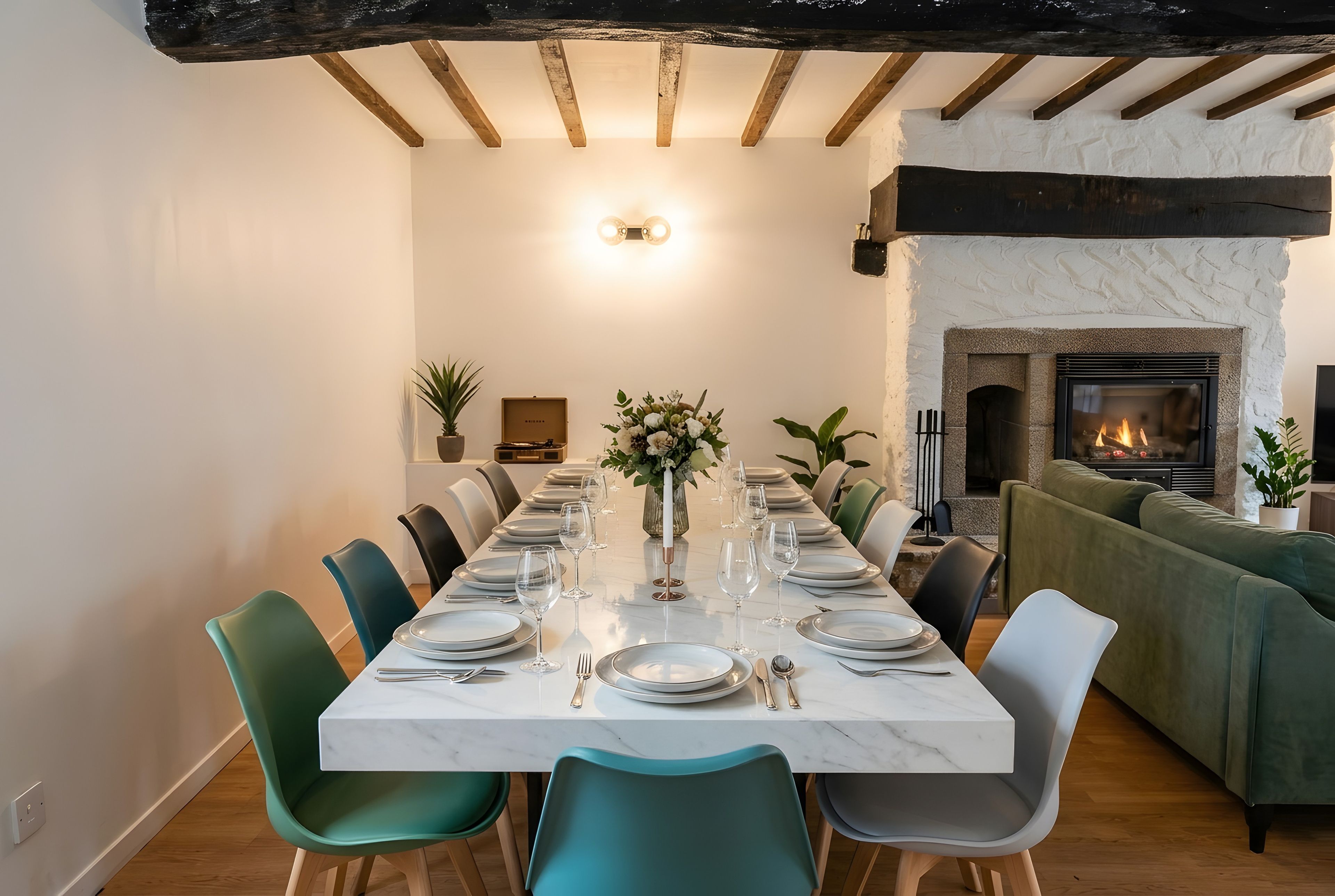 A dining room with a long table set for six, surrounded by teal chairs, a white marble table, and a fireplace in the background.