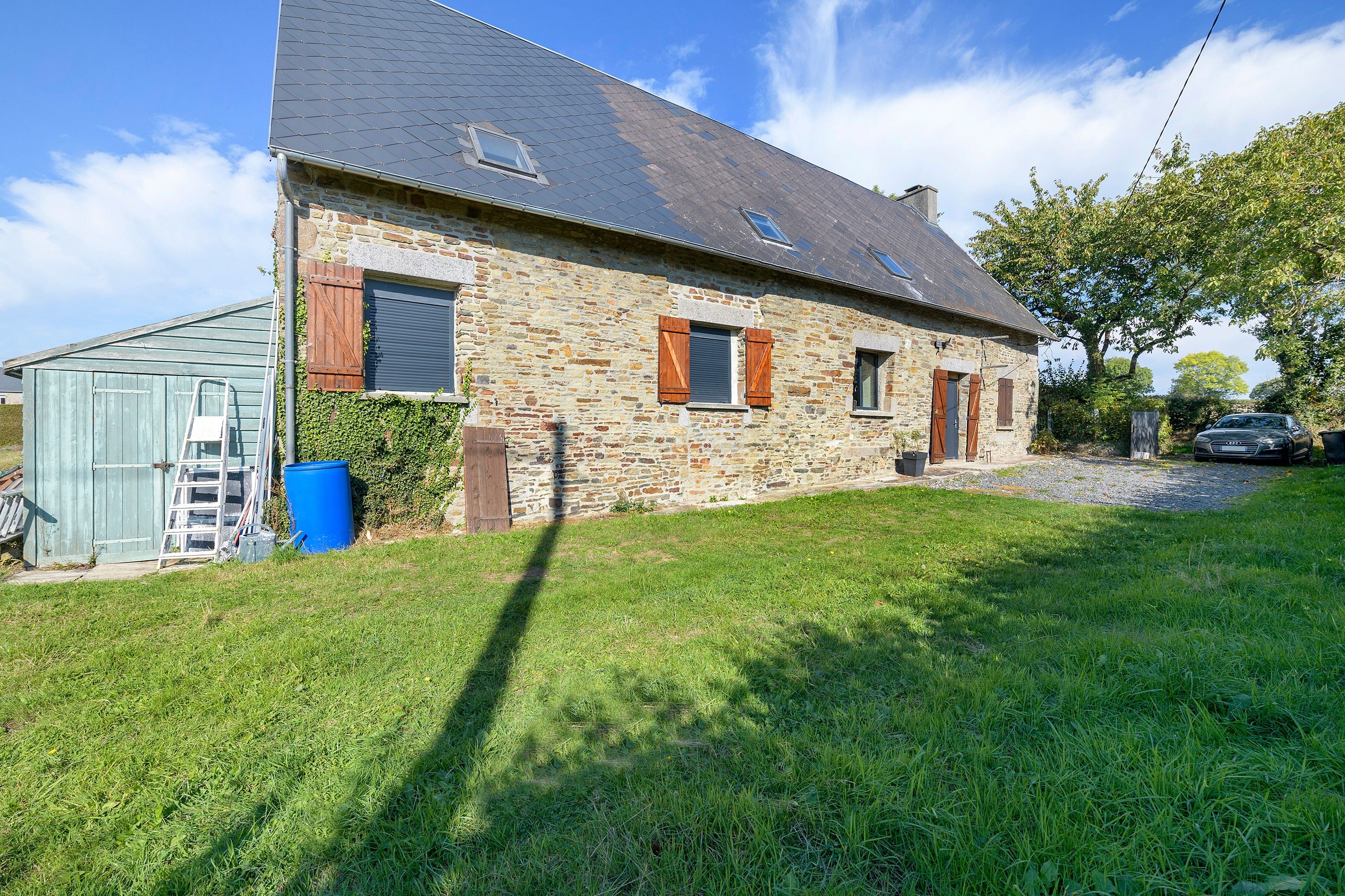 A stone house with a sloped roof, a small attached shed, and a car parked in the driveway.