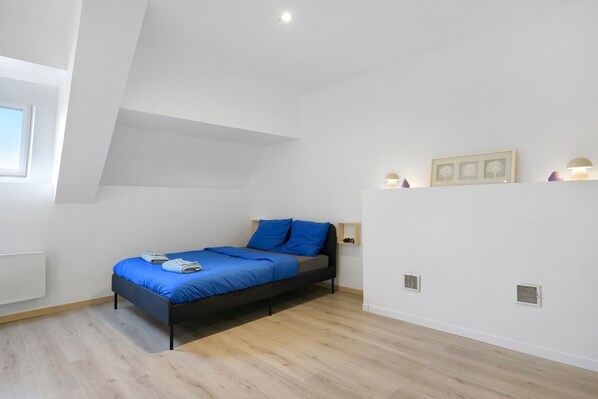 A bedroom with a bed, a window, a wall-mounted shelf with decorative items, and a white ceiling.