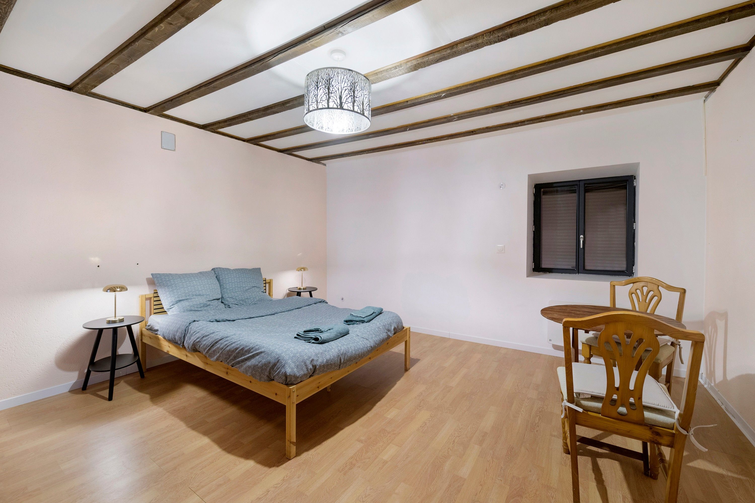 Ein Schlafzimmer mit einem hölzernen Bett, einem kleinen Tisch, einem Stuhl, einer Lampe und einem Fenster.