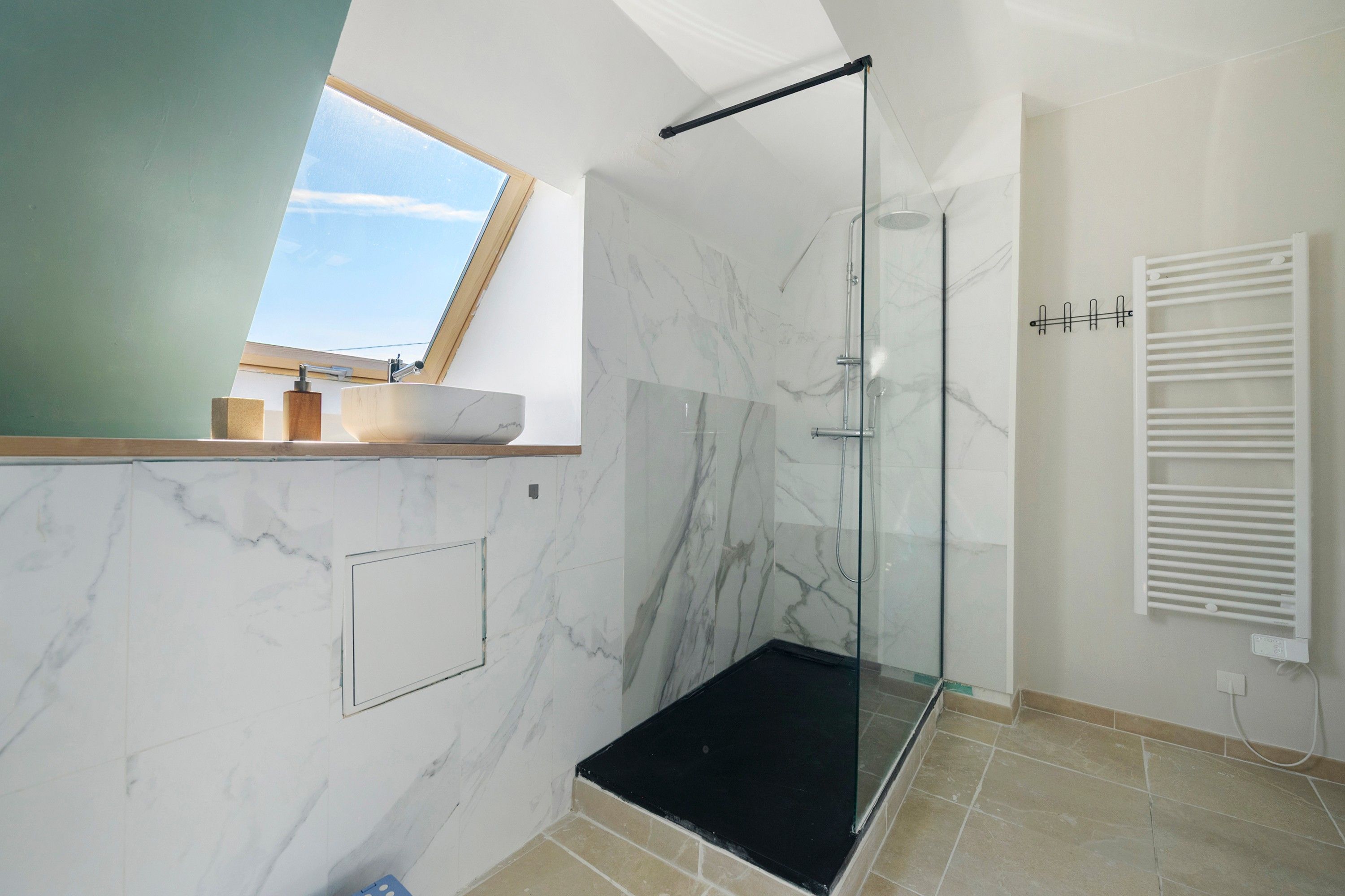 Une salle de bain moderne avec une douche à l’italienne en verre, un lavabo blanc et une fenêtre laissant entrer la lumière naturelle.