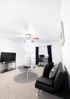 Un salon moderne doté d’un ventilateur de plafond, d’un téléviseur à écran plat, d’une table basse en verre, d’un canapé en cuir noir et d’un bureau avec un ordinateur.