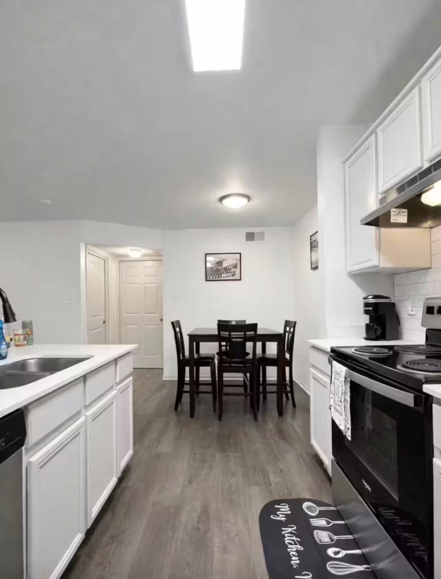 A kitchen with white cabinets, a dining table with chairs, a black stove, and a black rug with kitchen utensils.