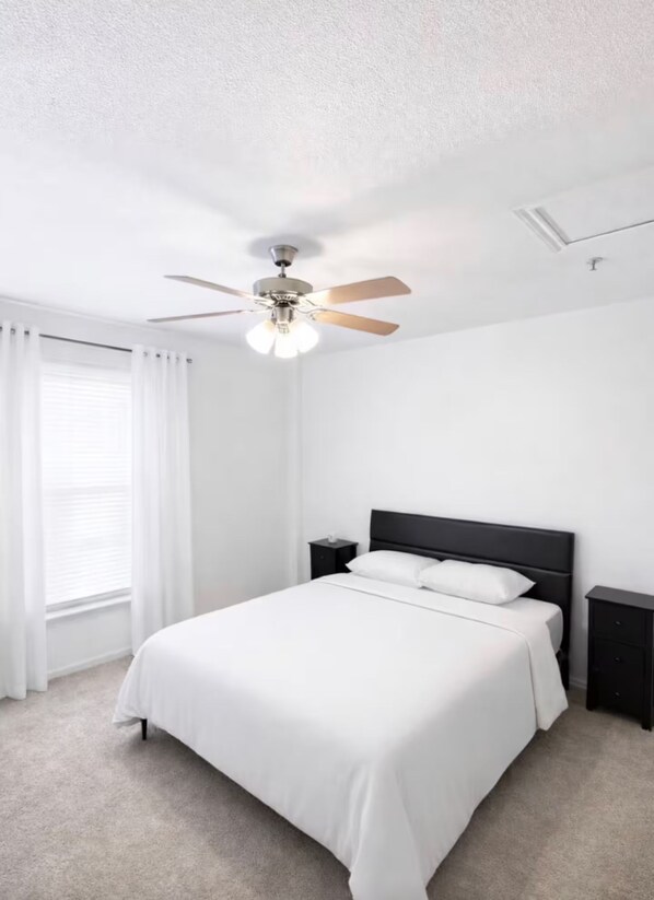 Une chambre à coucher avec un lit, un ventilateur de plafond, une fenêtre avec des rideaux et une table de chevet.