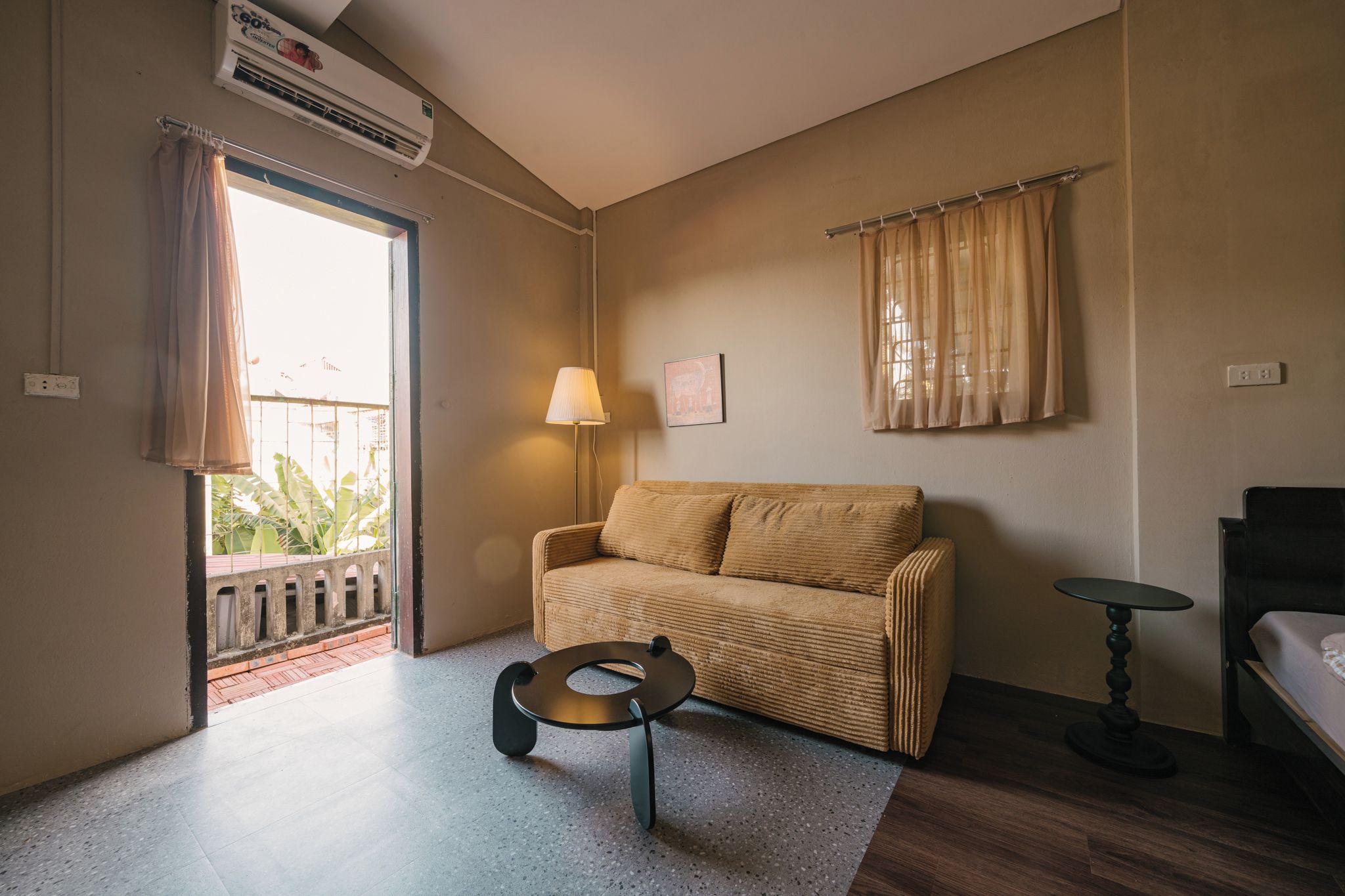 Une pièce comprenant un canapé, une petite table, une lampe et un lit. Il y a une fenêtre avec des rideaux et un appareil de climatisation installé au-dessus.