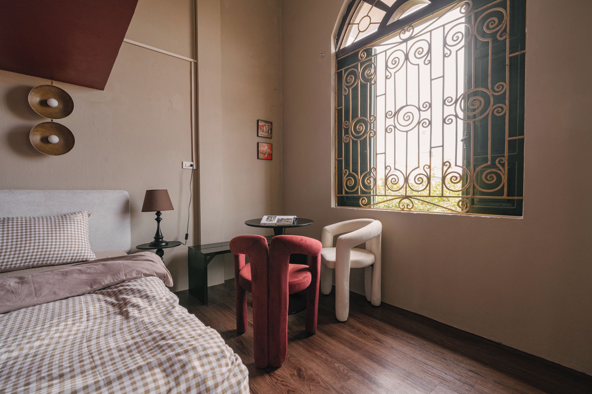 A bedroom with a bed, a desk, a chair, a window with decorative iron bars, and a wall-mounted lamp.