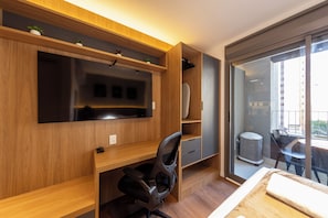 A modern office space with a large flat-screen TV, wooden shelving, a desk with a chair, and a sliding glass door leading to an outdoor area. - Valen by Bhomy (São Paulo)