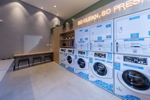 A laundry facility with multiple washing machines, a seating area, and a sign that reads 'SO CLEAN, SO FRESH'. - Valen by Bhomy (São Paulo)
