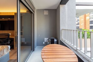 A modern apartment with a glass door leading to a bedroom, a small balcony with a wooden table, and a view of a building across the street. - Valen by Bhomy (São Paulo)
