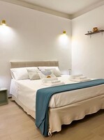 Una cama bien hecha con cabecera blanca, una manta azul y una mesita de noche con una lámpara.