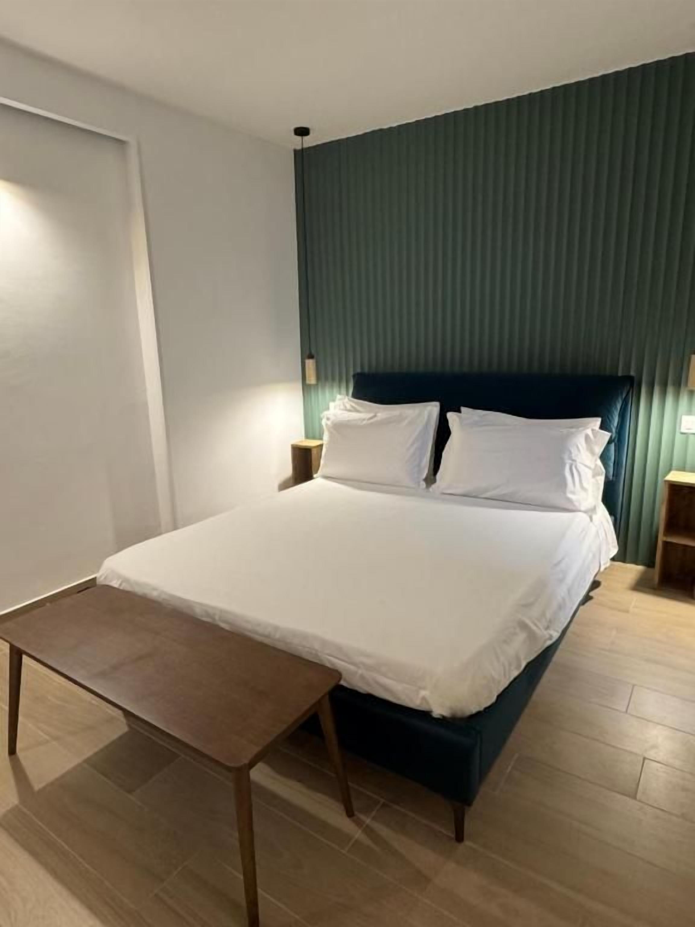 Una cama bien hecha con un cabecero oscuro, una pequeña mesa de madera y dos lámparas de noche.