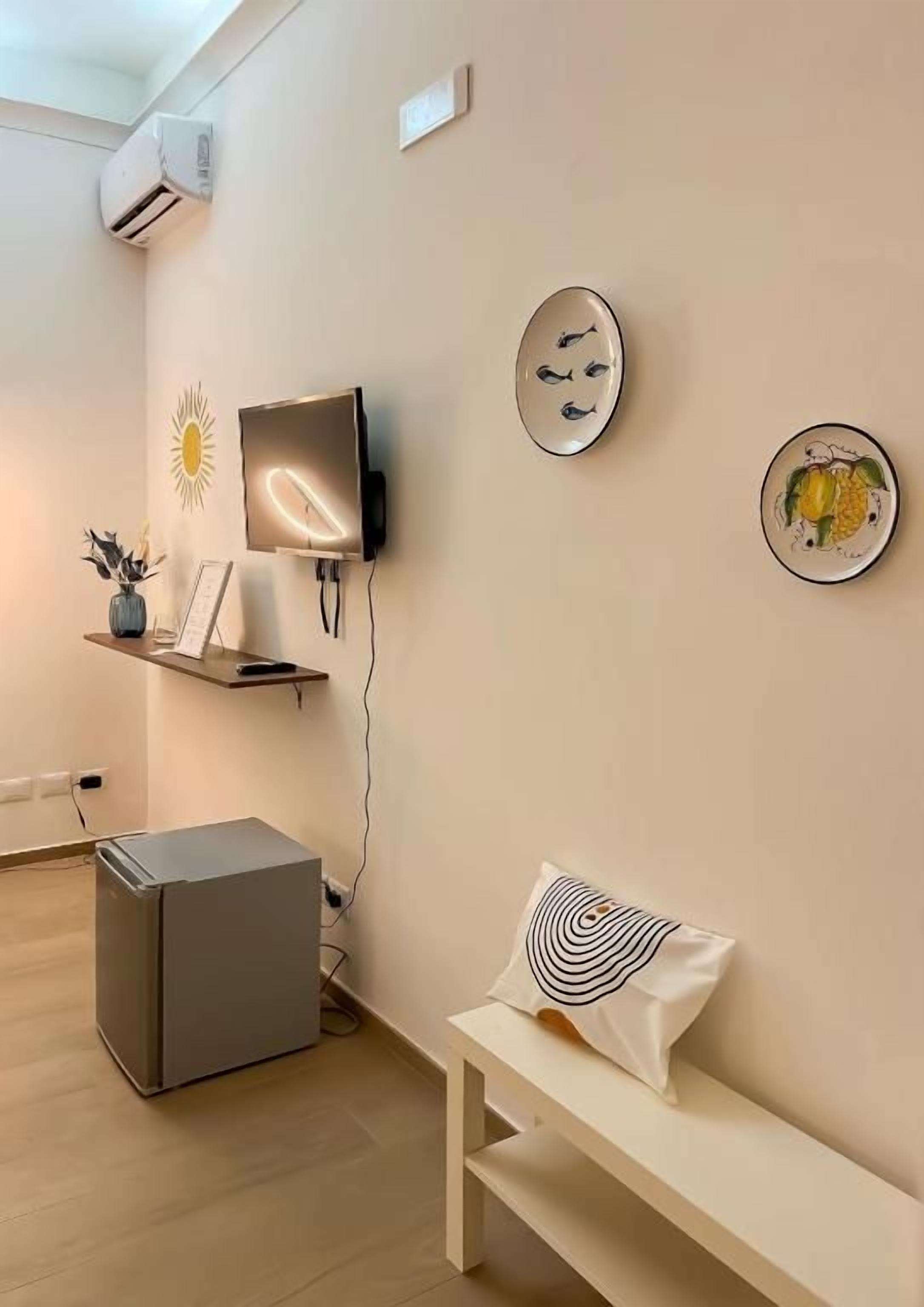 A minimalist room with a flat-screen TV, a small refrigerator, a bench, and wall-mounted decor.