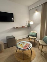 A modern living room with a flat-screen TV, a round table with a colorful floral tablecloth, a sofa, a small side table, and a lamp.