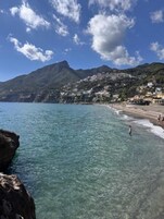 A coastal beach with clear water, a rocky shoreline, and buildings on a hillside.