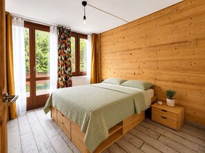 A bedroom with a wooden bed, a nightstand, and a view of greenery outside. - Apartment at the slopes - 8 guests (La Plagne)