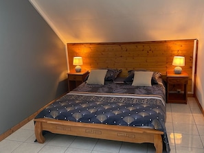 Ein Schlafzimmer mit Holz-Kopfteil, zwei Nachttischen mit Lampen, einem Bett mit gemusterten Betttextilien und einer kleinen Holzbank am Fußende.