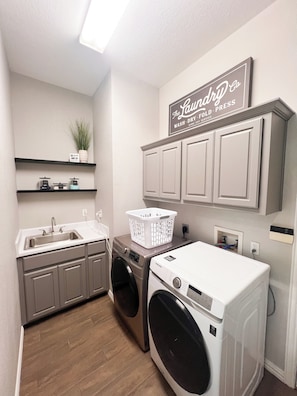 A laundry room with a washing machine, a dryer, a sink, and a shelf with a plant. - Stunning Poolside Retreat w/ Separate Pool House (Irving)